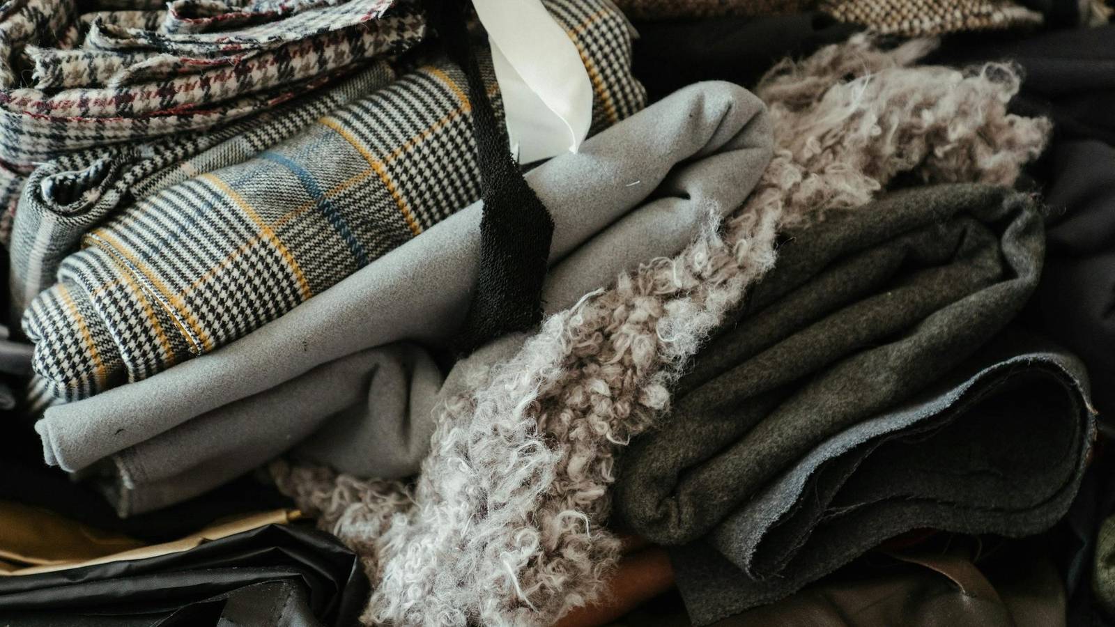 Folded lengths of wool cloth piled together: a black and white check, a plain grey, a dark green and a curly bouclé wool.